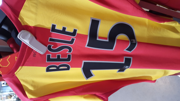 Ein roter und gelber Fußballtrikot mit Text und Nummern darauf, das an einer Stange in einem Geschäft hängt, zusammen mit anderen Gegenständen und einem sichtbaren Boden im Hintergrund.