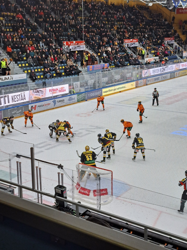 Gruppe von Menschen, die Hockey auf einem Eisring mit einem Netz und einer umgebenden Absperrung spielen, Zuschauern im Hintergrund, sichtbare Banner und Überdachungsbeleuchtung.