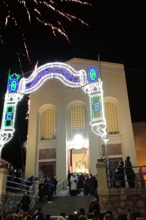Eine Gruppe von Menschen, die Silvester vor einem Gebäude mit einem beleuchteten Bogengang feiern, das mit Treppen und Geländern sowie Topfpflanzen geschmückt ist und unter einem Feuerwerkhimmel steht.