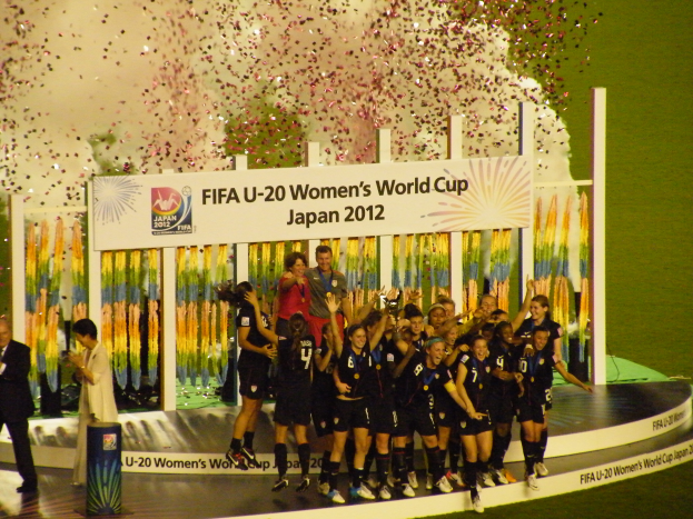 Eine Gruppe von Frauenfußballspielerinnen in schwarzen Uniformen steht auf einer Bühne, feiert ihren Sieg und ist von einer Menge mit Kameras umgeben.