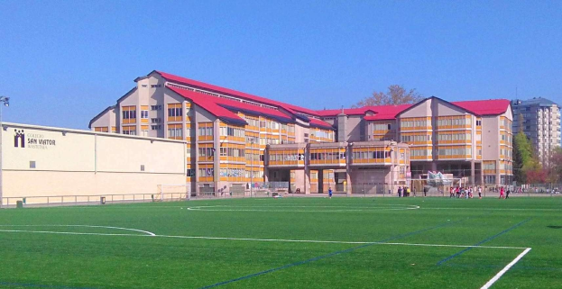 Ein Fußballfeld mit einem großen Gebäude im Hintergrund, das als San Visitor Academy beschriftet ist, umgeben von Bäumen und Laternenmasten, einige Menschen auf dem Feld und der Himmel darüber sichtbar.