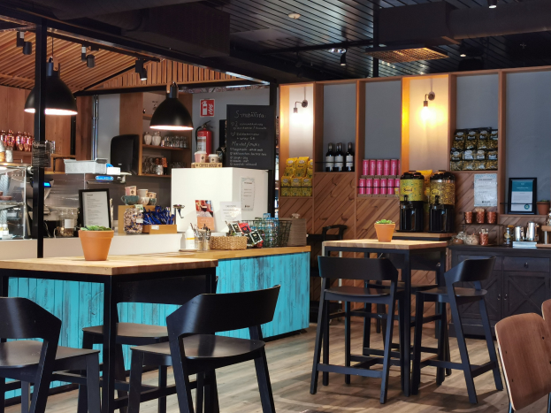 Innenraum eines Restaurants mit Holztischen, Stühlen, einem Topf mit einer Pflanze, einer Schiefertafel, einem Kühlschrank, einem Feuerlöscher, Regalen mit Flaschen, Schränken mit Gegenständen, einem Rahmenfoto an der Wand und Deckenleuchten.