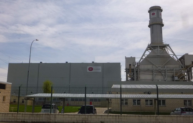 Ein großes Gebäude mit einem zentralen Turm, umgeben von einem Zaun, mit Fahrzeugen auf der benachbarten Straße, Masten, Lampen, Drähten, Bäumen und Gras im Vordergrund und einem bewölkten Himmel im Hintergrund, das eine neue Energiespeicheranlage darstellt.