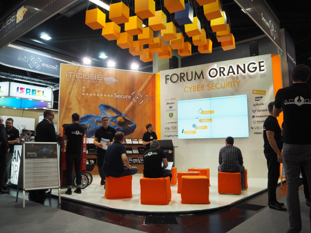 Eine Gruppe von Menschen an einem Messestand für IT-Cube Systems Forum Orange Cyber Security, mit sitzenden und stehenden Teilnehmern, einer Tafel mit Text, einem Bildschirm, Regalen mit Gegenständen, Schildern, Deckenleuchten und Lampen.