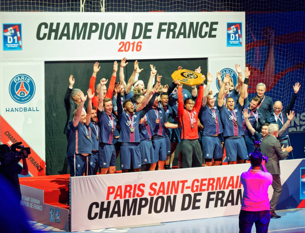 Gruppe von Männern, die einen Pokal auf einer Bühne halten, mit "Champion de France 2016" im Hintergrund, eine Person filmt die Feier.
