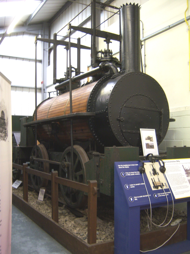 Alte Dampflokomotive in einem Museum mit Informationsschildern, einem Zaun, Steinen, einer Fahne, einer Wand mit Rohren und Deckenleuchten ausgestellt.