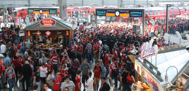 Eine große Menschenmenge, die durch einen belebten Bahnhof geht, mit einem Zug auf den Eisenbahnschienen im Hintergrund, mit Geschäften, Texttafeln, Pfählen, Lampen und anderen Bahnhofsobjekten.