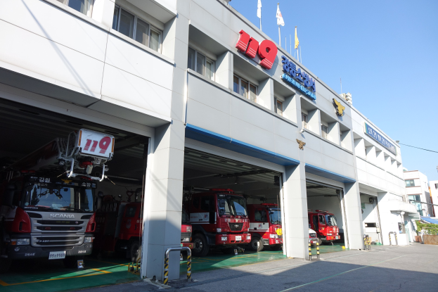 Feuerwache mit mehreren Feuerwehren vor dem Gebäude geparkt, mit Fenstern, Säulen, Namensschildern und Flaggen im Vordergrund, vor anderen Gebäuden unter einem klaren blauen Himmel.