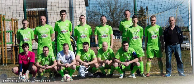 Eine Gruppe von Männern in grünen T-Shirts steht auf einem Fußballfeld mit einem Ball und einem Torpfosten, einige hocken, mit Bäumen, Gebäuden und Fahrzeugen im Hintergrund.