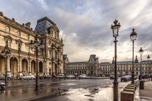 Außenansicht des Louvre-Museums in Paris, das seine charakteristische Architektur, Straßenlaternen, Lichter, Fahrzeuge, Fußgänger auf dem Gehweg und einen bewölkten Himmel zeigt.