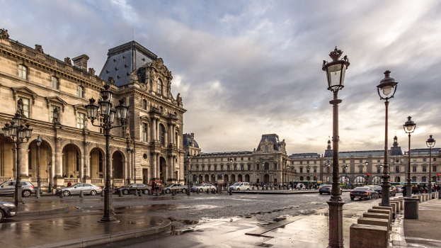 Außenansicht des Louvre-Museums in Paris, das seine charakteristische Architektur, Straßenlaternen, Lichter, Fahrzeuge, Fußgänger auf dem Gehweg und einen bewölkten Himmel zeigt.