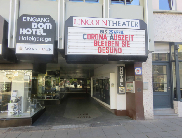 Außenansicht des Lincoln Theaters in Berlin mit Glasfenstern, -türen und einer Texttafel sowie inneren Elementen, die eine lebendige städtische Szenerie suggerieren.