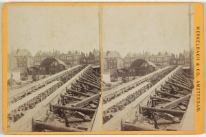 Ein altes Schwarz-Weiß-Foto einer Bahntrasse mit Gebäuden im Hintergrund, Text auf der rechten Seite, der 'Merkelbach & Co. Amsterdam' lautet, und verstreute hölzerne Gegenstände im Vordergrund.