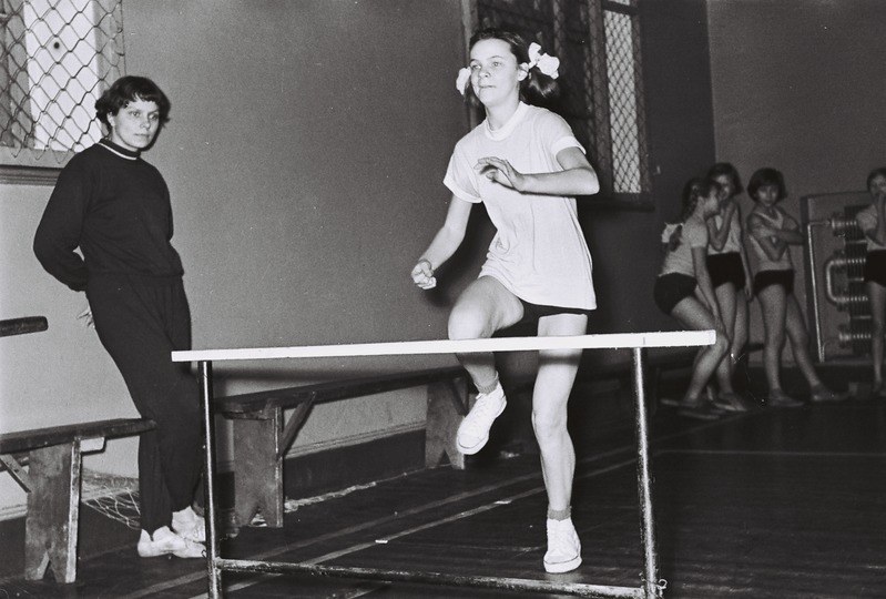 Schwarzes und weißes Foto einer Frau, die über ein Hindernis in einer Halle springt, mit Zuschauern im Hintergrund.