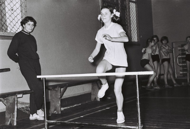 Schwarzes und weißes Foto einer Frau, die über ein Hindernis in einer Halle springt, mit Zuschauern im Hintergrund.
