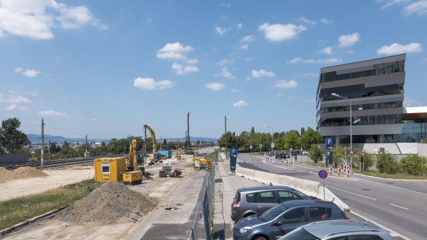 Eine Baustelle mit geparkten Autos, Gebäuden, Straßenlaternen, Straßenschildern, Absperrpoller, Bäumen, Pflanzen, Gras, Sand, Kränen, Bahnschienen, Strommasten mit Kabeln, Hügeln und einem bewölkten Himmel im Hintergrund.