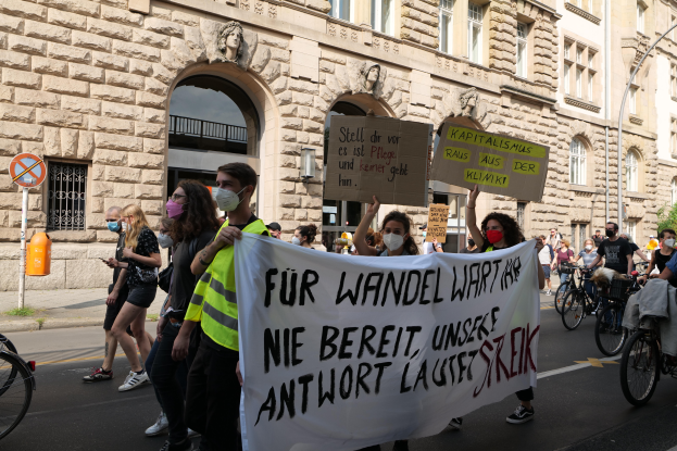 Eine Gruppe von Menschen marschiert auf einer Straße in Berlin, mit Schildern und Transparenten, einige fahren Fahrräder, vor einem Gebäude mit Fenstern, Überdachungen, Pfeilern, Skulpturen und Bäumen.