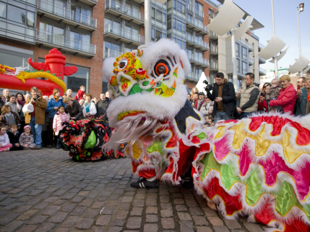 Ein lebendiges chinesisches Neujahrsfest in Amsterdam mit einer Löwen-Tanz-Performance vor einer Zuschauermenge, darunter einige mit Kameras, vor einer Kulisse aus Gebäuden, Laternenmasten und einem klaren blauen Himmel.