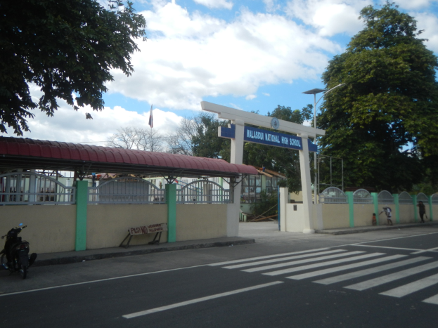 Einfahrt zu einer Schule mit einem vor der Tür geparkten Motorrad, einer Straße mit weißen Linien und Zebrastreifen, einer Mauer mit einem Schuppen, einem Schild, Laternenpfählen, Bäumen, einer Fahne auf einem Mast und einem bewölkten Himmel.