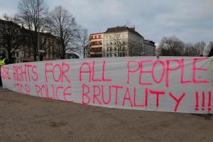 Eine Gruppe von Menschen, die auf dem Boden stehen und eine Fahne halten, auf der steht "Rechte für alle Menschen Stoppt Polizeigewalt", mit einem Straßenschild, einem Schild, Bäumen, Gebäuden mit Fenstern und einem bewölkten Himmel im Hintergrund.