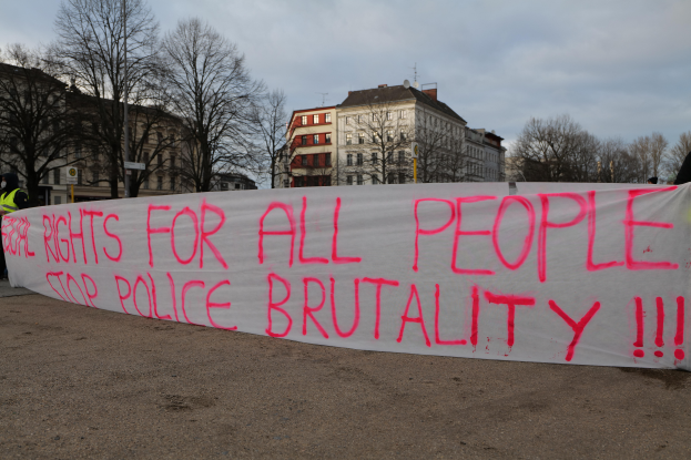 Eine Gruppe von Menschen, die auf dem Boden stehen und eine Fahne halten, auf der steht "Rechte für alle Menschen Stoppt Polizeigewalt", mit einem Straßenschild, einem Schild, Bäumen, Gebäuden mit Fenstern und einem bewölkten Himmel im Hintergrund.