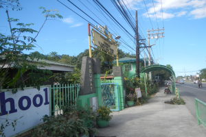Schuleingang mit einem "Schule"-Schild, Fahrzeuge auf der Straße, ein Torbogen, ein Tor, eine Namenskarte, Pflanzen, Bäume, Strommasten mit Drähten und eine bewölkte Himmel im Hintergrund.