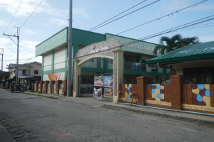 Ein Schulgebäude mit einem grünen Dach an einer Straßenecke, umgeben von Fahrzeugen, Werbetafeln, Strommasten, Bäumen und einem bewölkten Himmel.