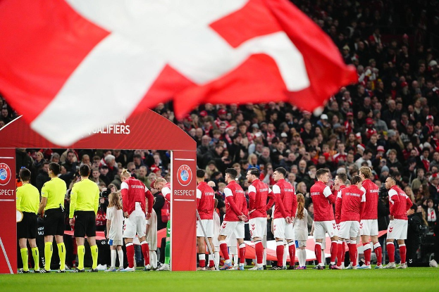 Eine Gruppe von Menschen auf einem Fuballfeld mit einer roten und weißen Flagge im Vordergrund, einem Bogen mit der Aufschrift "Bayern München vs Bayern München Wetten & Vorschau" im Hintergrund und einer großen Menge im Stadion.