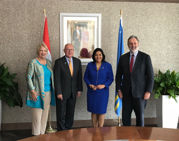 Eine Gruppe strahlender Menschen um einen Tisch mit Papieren herumstehend, umgeben von Fahnen, Topfpflanzen und einem Foto auf dem Wand, die die Unterzeichnung eines Memorandums von Verständnis zwischen den Vereinigten Staaten und Kanada feiern.