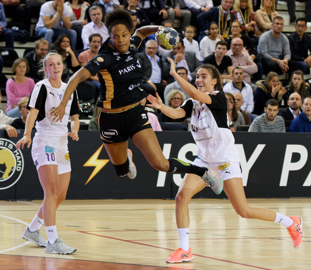Eine Gruppe von Frauen, die Handball auf einem Platz spielen, mit einem Ball in der Mitte, Zuschauern im Hintergrund und einem Banner mit Text und Logos.