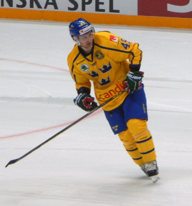 Eishockeyspieler in Bewegung auf dem Eis mit Helm, Handschuhen und Schlittschuhen, der einen Hockeystock hält, mit einem Schild im Hintergrund mit Text.