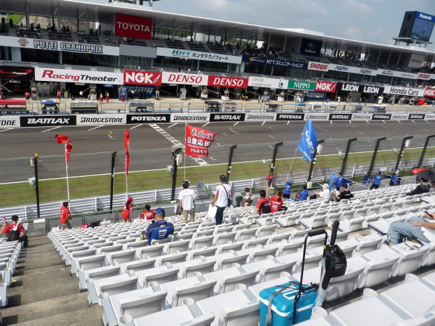 Eine Gruppe von Menschen sitzt auf der Tribüne einer Rennstrecke, umgeben von Fahnen, Schildern, Geländern und Stufen, unter einem bewölkten Himmel.