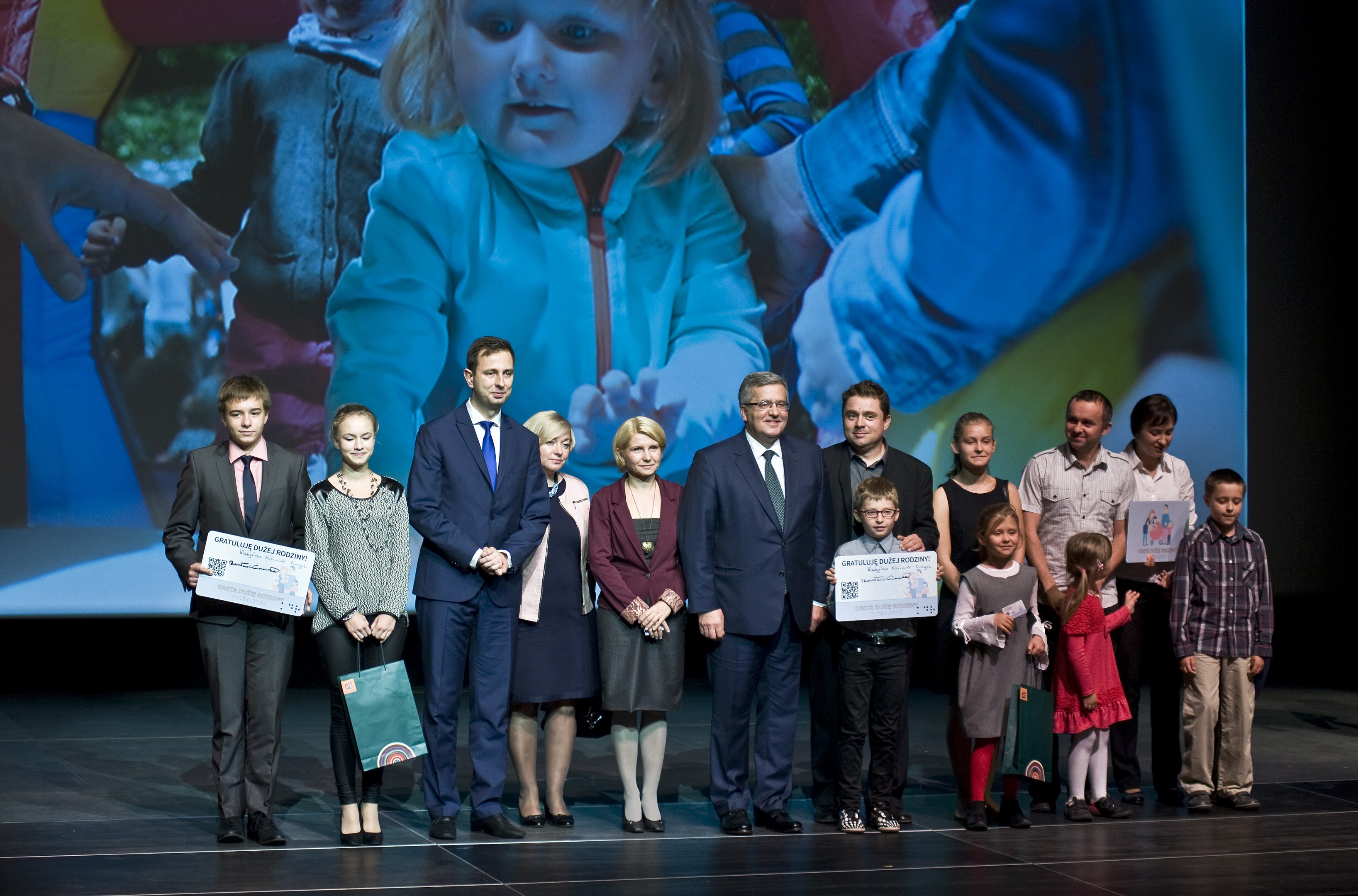 Gruppe von Menschen auf einer Bühne mit Taschen und Urkunden, im Hintergrund ein Bildschirm mit dem Bild eines Kindes und anderen