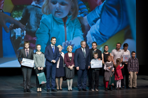 Gruppe von Menschen auf einer Bühne mit Taschen und Urkunden, im Hintergrund ein Bildschirm mit dem Bild eines Kindes und anderen