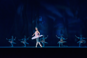 Eine Ballerina in rosafarbenem Tutu und Spitzenschuhen tanzt anmutig auf der Bühne, umgeben von anderen ähnlich gekleideten Tänzern, mit einem dunklen Hintergrund, der eine dramatische Atmosphäre schafft.