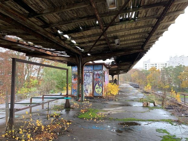 Ein verlassenes Bahnhofsgebäude mit Graffiti, umgeben von Bäumen, Gebäuden, Geländern, Pfählen, Laternen und einem Schuppen, mit trockenem Laub und Gras auf dem Boden und dem Himmel im Hintergrund.