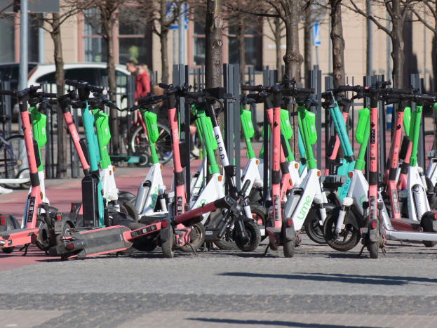 Eine Reihe von Elektro-Rollern, die an einer Straße geparkt sind, mit einer Person im Vordergrund und unscharfen urbanen Elementen im Hintergrund.