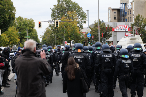 Eine Gruppe von Menschen, die eine Straße entlanggehen, mit Polizeibeamten in der Nähe, einige tragen Helme und tragen Taschen oder Kameras, mit Fahrzeugen, Verkehrszeichen, Schildern, Bäumen, Gebäuden und einem klaren Himmel im Hintergrund.