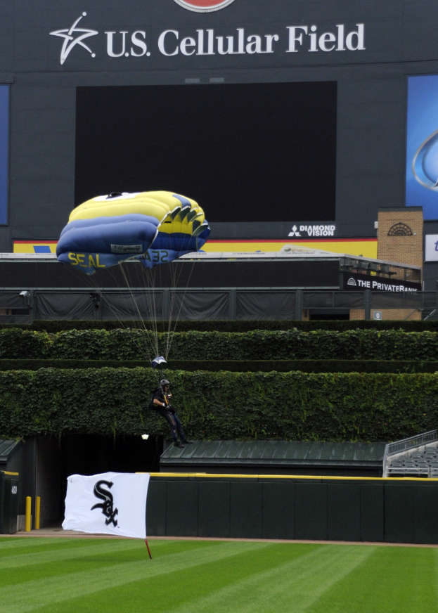 Eine Person, die über ein Baseballfeld mit einem Fallschirm gleitet, umgeben von Grünflächen, einem Banner, Tafeln mit Text und Logos und einem Gebäude im Hintergrund.