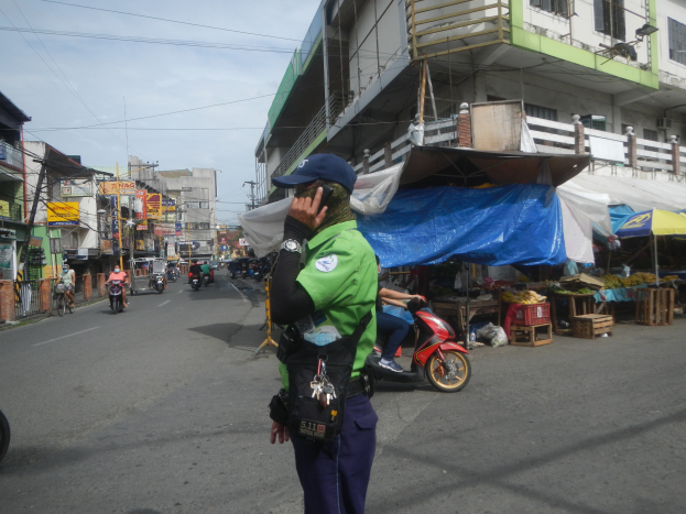 Ein Polizist in grüner und blauer Uniform und blauer Mütze steht an der Straße und spricht in ein Handy, während Autos im Hintergrund fahren und Stände mit verschiedenen Gegenständen rechts von ihm sind.