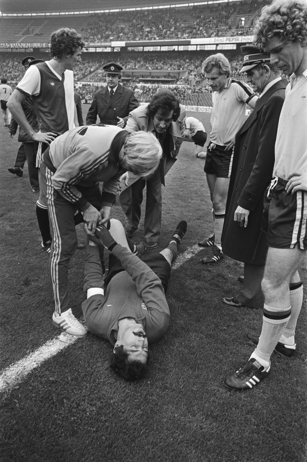 Schwarzes Bild eines Fußballspielers, der auf dem Boden liegt und von einer Gruppe Männer umgeben ist, mit einer Menge im Stadionhintergrund.