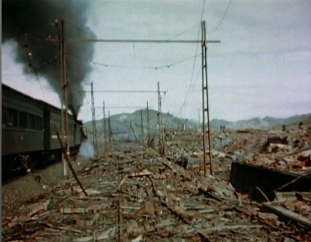 Ein Zug fährt durch ein beschädigt Gebiet mit Rauch, der aus ihm aufsteigt, umgeben von Masten und Dröhnen, mit Hügeln und einem klaren Himmel im Hintergrund und verstreutem Schutt auf dem Boden.