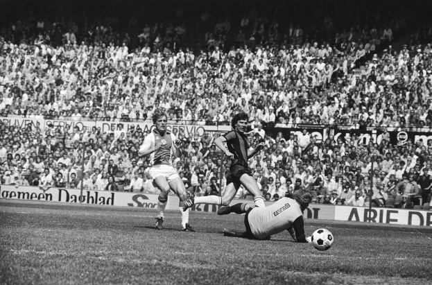 Schwarzes und weißes Foto von Füßballspielern im Einsatz auf einem Feld mit Zuschauern im Hintergrund und einer Bundesliga 1978-79 Ajax 1-1 Hannover 96-Banner.