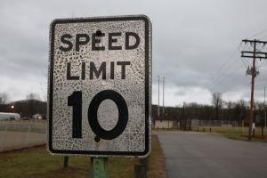Tempobegrenzungsschild am Straßenrand mit Gras, Strommasten, Drähten, einem Zaun, Häusern, Bäumen und einem bewölkten Himmel im Hintergrund.