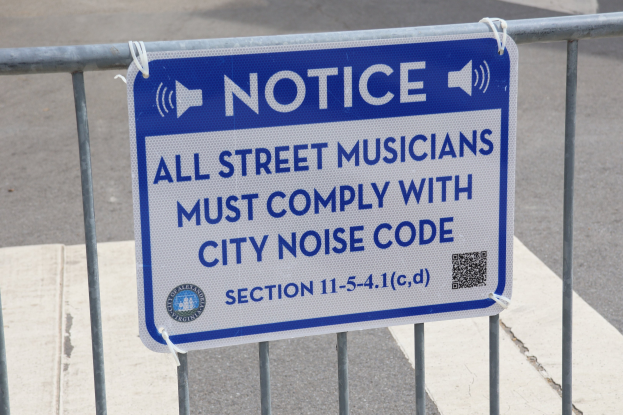 Anschlagtafel mit einem Schild, auf dem "Hinweis: Alle Straßenmusiker müssen die städtische Lärmvorschrift einhalten" steht, an einem Zaun befestigt, mit einer Straße im Hintergrund.