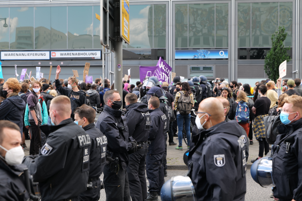 Eine große Gruppe von Menschen vor einem Gebäude stehend, einige halten Schilder und tragen Helme, mit einem Pfahl mit Schildertafel im Vordergrund und einem Baum im Hintergrund, scheinbar protestierend.