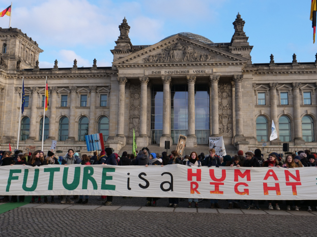 Eine Gruppe von Menschen hält ein Transparent mit der Aufschrift "Zukunft ist ein Menschenrecht" vor dem Reichstaggebäude in Berlin, Deutschland, mit sichtbaren Fenstern, Säulen und Bögen, umgeben von Fahnenmasten, unter einem bewölkten Himmel.