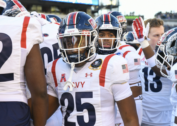 Gruppe von Männern in Fußballausrüstung auf einem Feld mit Helmen und Handschuhen, Hintergrund zeigt unscharfe Zuschauer, ein Anzeigebrett mit der Aufschrift "Auburn vs Virginia Tech - 11/28/15 College Football Pick, Odds, and Prediction" und Himmel.