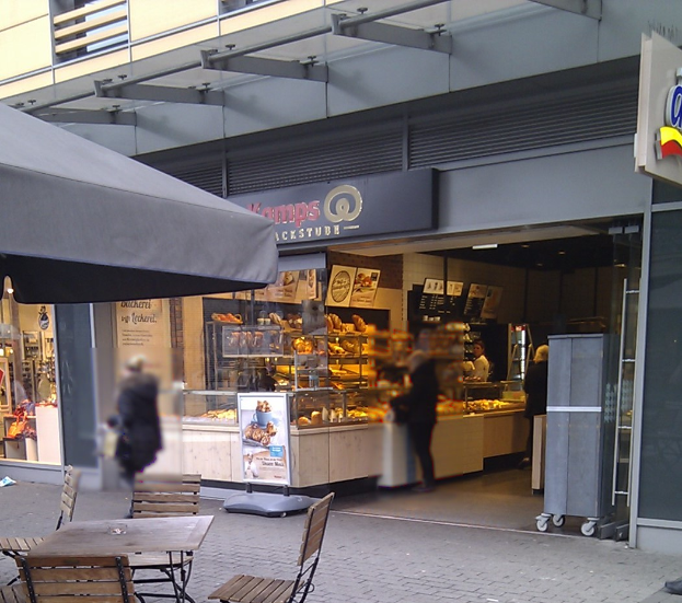 An outdoor restaurant with tables, chairs, and an umbrella, surrounded by pedestrians and a building in the background.