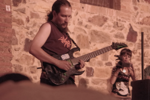 Ein bärtiger Mann spielt eine Gitarre auf der Bühne, rechts von ihm hält eine Frau ein Mikrofon und im Hintergrund ist eine Menschenmenge zu sehen.